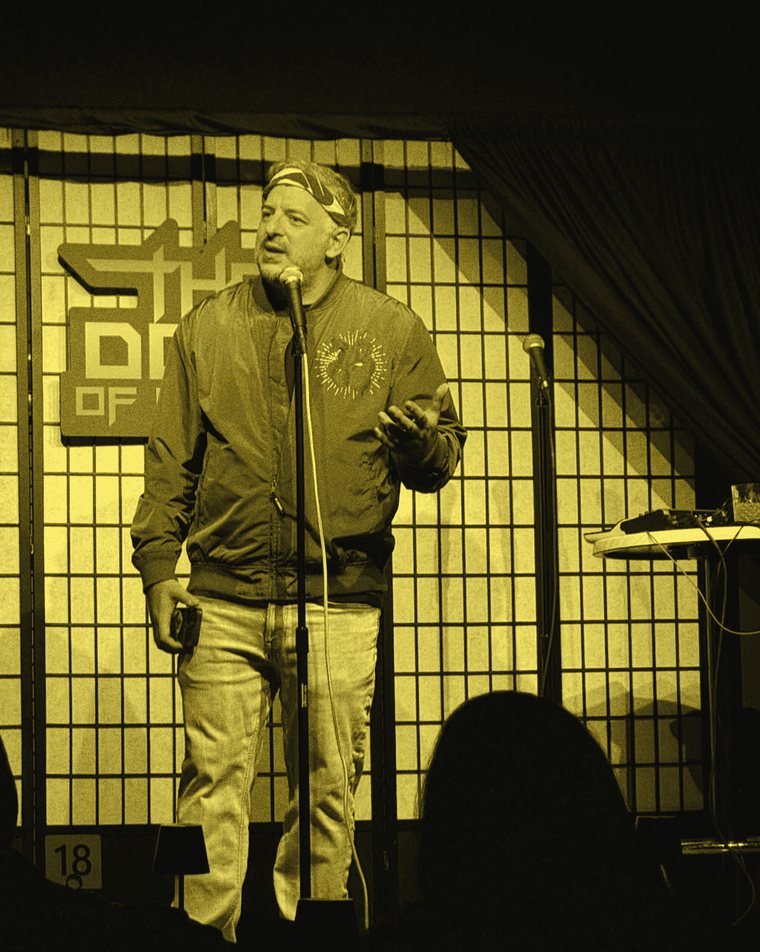 A performer in a red bandana and gray jacket speaking at a microphone, one hand open