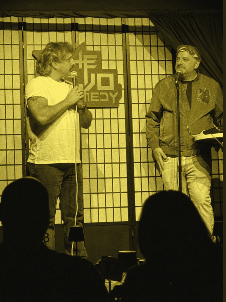 Two performers on stage looking at one another across a microphone stand