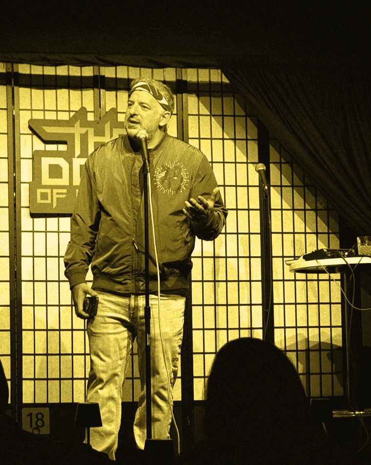 A performer at a microphone in front of the Dojo of Comedy sign, one hand open mid-gesture