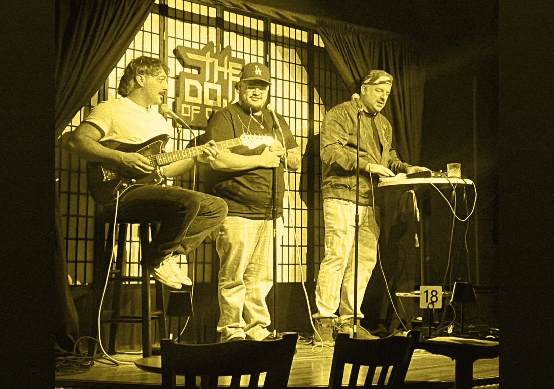 A guitarist on a stool, a performer holding a microphone, and a performer at a table of instruments, on stage at the Dojo of Comedy