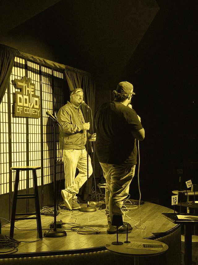 A comedian with his back to the camera speaking to a performer in a red bandana on stage