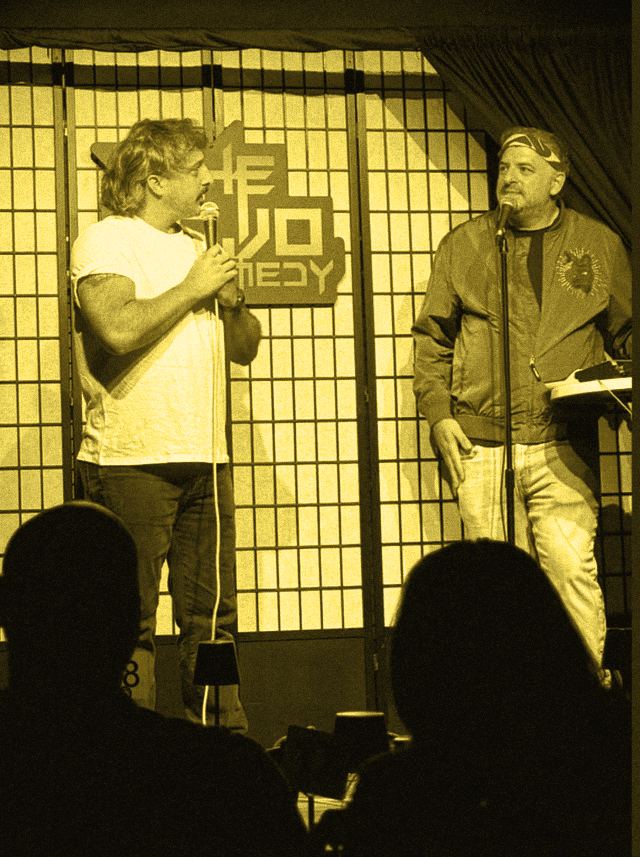 Two performers on stage regarding one another across a microphone stand